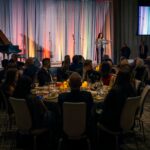 A formal event with guests seated at round tables, listening to a speaker on stage in front of a colorful curtain backdrop, with a grand piano nearby.