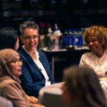 Guests seated at a round table, engaged in conversation and smiling at a formal event. A beverage station with bottled water and ice buckets is visible in the background.