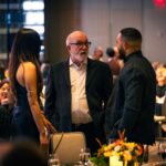 An older man with glasses and a white beard engaged in conversation with a younger man and woman at a formal dinner event. The elegantly decorated venue features candlelit tables with floral centerpieces.