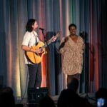 A musician playing an acoustic guitar and a singer stand on a stage with colorful draped curtains, performing in front of an audience.