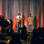 A musical trio performs on stage in front of an audience, featuring a pianist, a guitarist, and a vocalist. The backdrop is draped with colorful lighting.