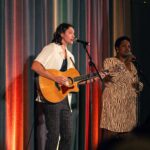 A male guitarist and a female singer perform on stage, both singing into microphones against a colorful draped backdrop. The guitarist wears a white shirt and plaid pants, while the singer wears a patterned dress.