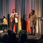 A musical trio performs on stage; a guitarist and a singer stand at microphones while a pianist plays in the background, with a colorful draped backdrop behind them.