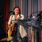 A musician with shoulder-length hair plays an acoustic guitar and sings into a microphone on stage. The background features colorful curtains, and a pianist is partially visible behind him.