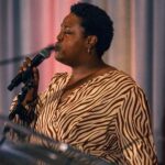 A singer passionately performs on stage, holding a microphone and wearing a zebra-patterned outfit, with a colorful draped backdrop behind them.