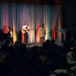 A live musical performance on stage featuring a guitarist and a vocalist, with a pianist accompanying them. The stage is lit with colorful curtains in the background, and an audience is seated in the foreground watching the performance.