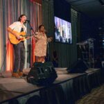 A guitarist and a singer perform on stage in front of microphones, with a large screen displaying their live performance in the background. The stage is decorated with warm lighting.