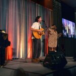A guitarist and a singer perform on a stage with a colorful draped backdrop, while a large screen displays their live performance to the audience.