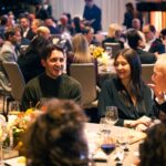 A lively dinner event with elegantly dressed guests engaged in conversation. A smiling man in a dark green sweater and a woman in a black dress share a moment of laughter at a candlelit table adorned with wine glasses and floral centerpieces.