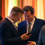 Two men in suits are smiling and looking at a smartphone together in a warmly lit setting with colorful vertical lights in the background.