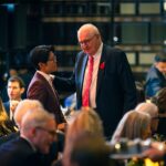 Two elegantly dressed individuals engaged in conversation at a formal event, with one placing a hand on the other's shoulder. The background shows a lively banquet setting with guests and warm lighting.