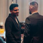 Two men engaged in conversation at a formal event, one wearing a black suit and the other in a dark sweater, with a softly lit background.