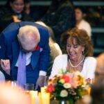 An elderly man in a blue suit with a purple tie leans down while speaking to a smiling elderly woman in a white outfit with pearl jewelry. A floral centerpiece and candles are on the table in front of them.