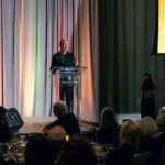A woman in a black outfit stands behind a clear podium at the Four Seasons Hotel, delivering a speech to a seated audience. The stage is lit with colorful drapery, and a woman in black stands in the background holding papers.