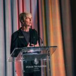 A person stands behind a clear podium at the Four Seasons Hotel Toronto, delivering a speech with a colorful draped backdrop behind them.