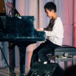 A young boy in a white long-sleeve shirt and black pants playing a grand piano on stage. He is focused, with his fingers on the keys, and a curtain backdrop is visible behind him.