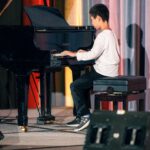 A young boy in a white long-sleeve shirt and black pants sits on a piano bench, playing a black Yamaha grand piano on a stage with colorful curtains in the background.