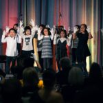 A group of children wearing white gloves performs on stage in front of an audience, raising their hands in unison. The stage is decorated with colorful curtains, and microphones are set up for the performance.