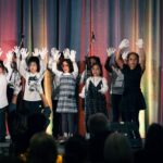 A group of children performing on stage, wearing white gloves and raising their hands in unison. A colorful curtain backdrop and microphones are visible behind them, with an audience watching in the foreground.