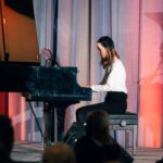A young woman in a white blouse and black pants playing a grand piano on stage. She is focused, with her fingers on the keys, and a curtain backdrop with red and gray lighting is visible behind her.
