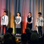 Four children stand on stage in front of microphones, dressed in formal and semi-formal attire, performing in front of an audience with a colorful draped backdrop behind them.