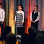 Two young girls stand at microphones on stage, singing in front of an audience, with a third child partially visible on the left. The backdrop is draped with colorful lighting.