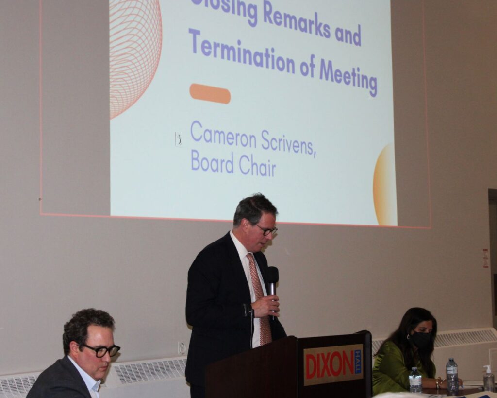 a-man-cameron-scrivens-is-giving-closing-remarks-at-a-meeting-while-standing-at-a-podium-labeled-dixon-hall-a-large-screen-behind-him-displays-his-name-and-role-as-board-chair-two-panelists-are-seated-at-a-table-beside-him-one-taking-notes-and-the-other-wearing-a-mask