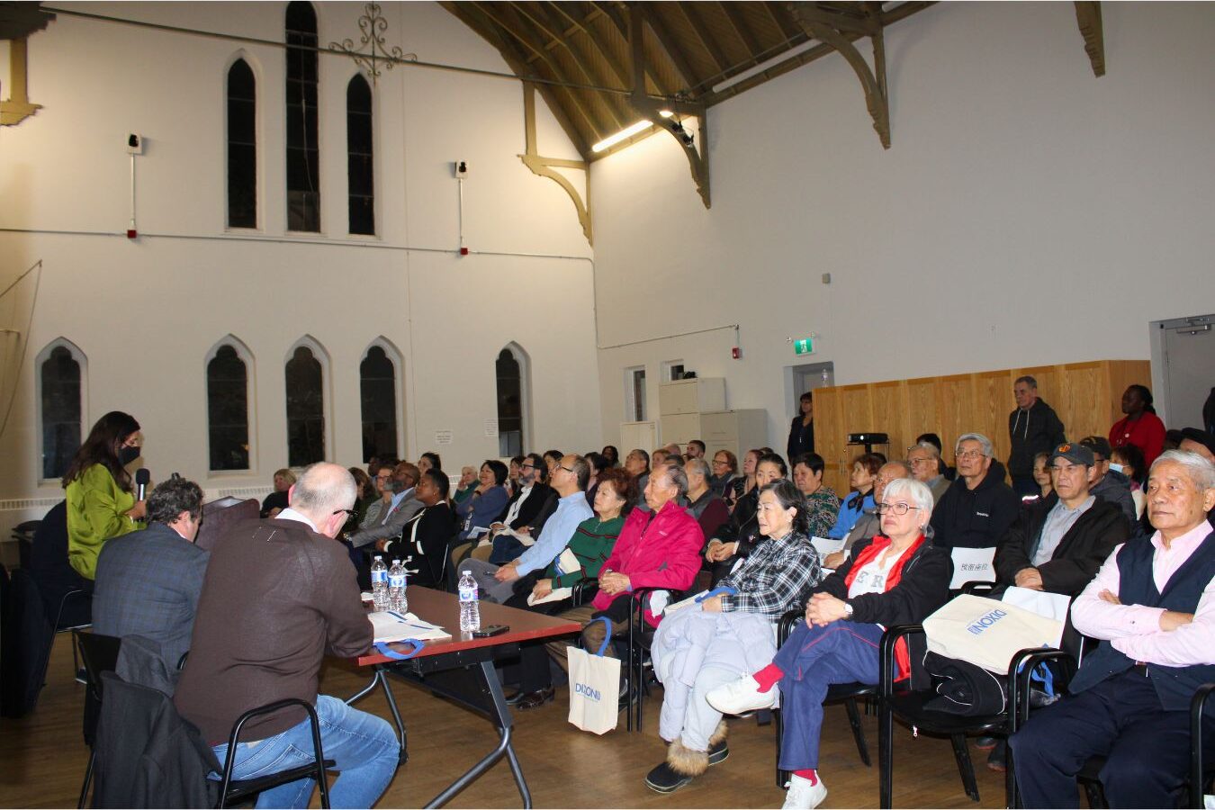 a-large-audience-is-seated-in-a-hall-attentively-listening-to-a-speaker-at-the-front-the-room-has-high-ceilings-with-arched-windows-and-panelists-are-seated-at-a-table-near-the-speaker-some-attendees-are-holding-tote-bags-labeled-dixon