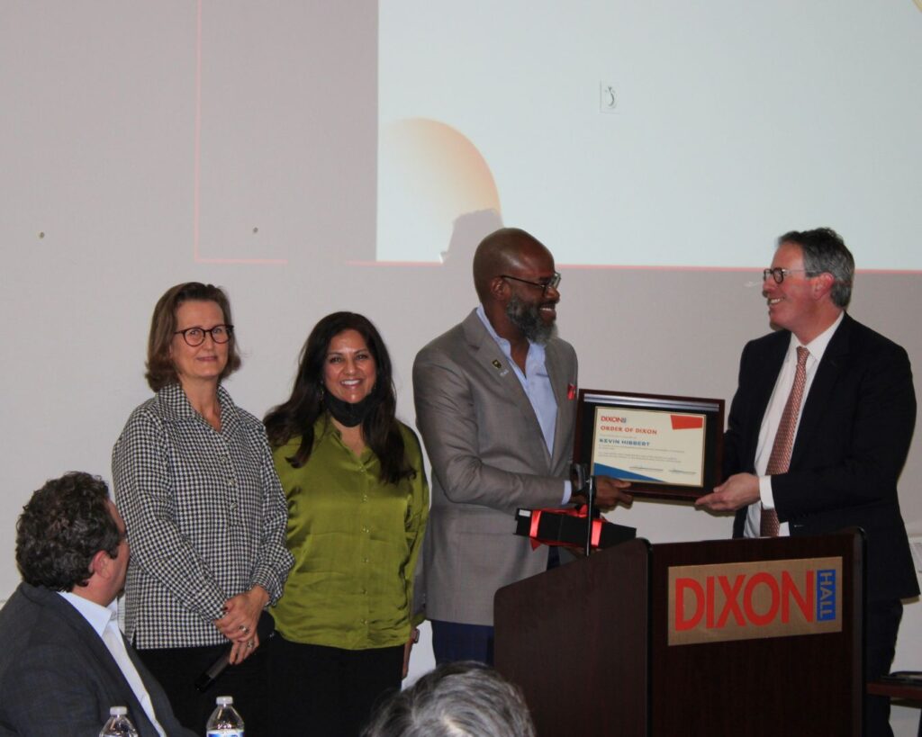 a-man-in-a-suit-is-presenting-a-framed-award-to-another-man-at-a-podium-labeled-dixon-hall-two-women-stand-beside-them-smiling-a-large-screen-in-the-background-displays-part-of-a-presentation