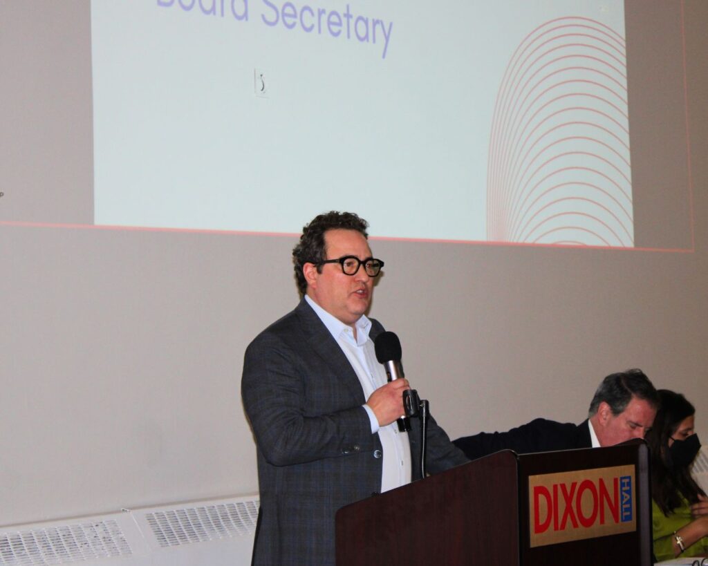 a-man-is-speaking-at-a-podium-labeled-dixon-hall-while-holding-a-microphone-a-large-screen-behind-him-displays-text-mentioning-board-secretary-two-panelists-are-seated-at-a-table-beside-him-one-taking-notes-and-another-wearing-a-mask