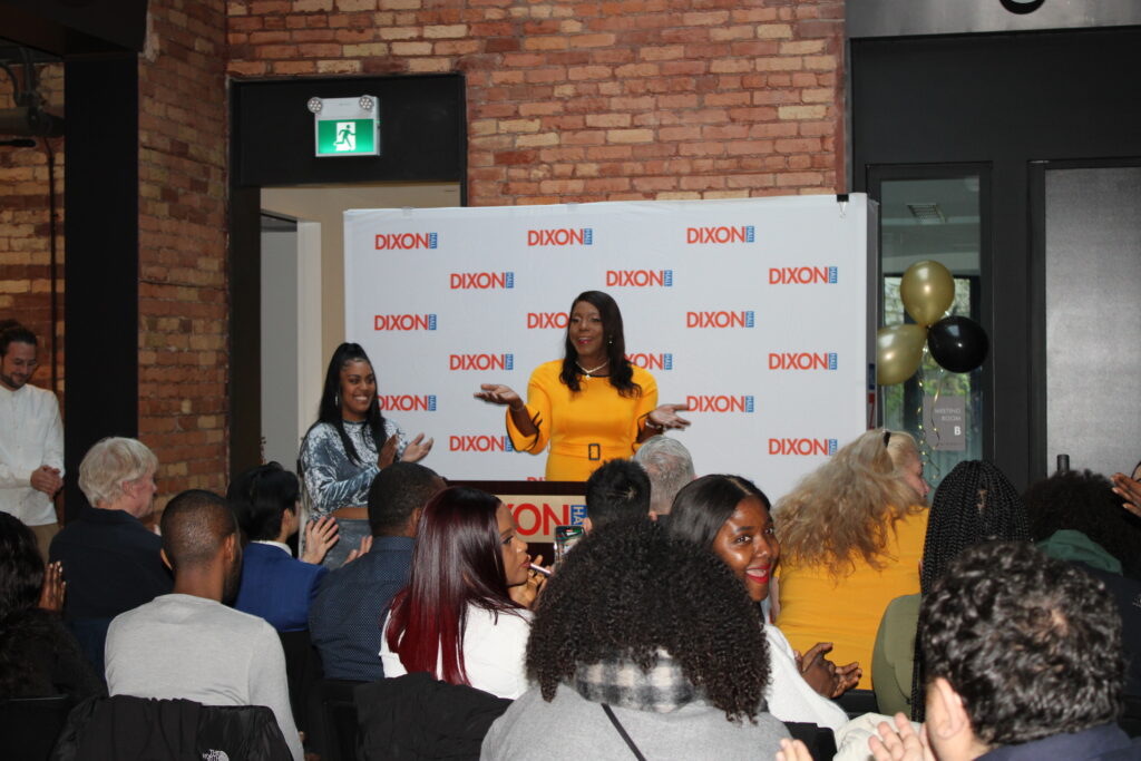 a-woman-in-a-yellow-dress-is-speaking-at-a-podium-labeled-dixon-hall-while-the-audience-applauds-another-woman-stands-beside-her-smiling-the-backdrop-features-the-dixon-hall-logo-and-black-and-gold-balloons-are-visible-in-the-background-