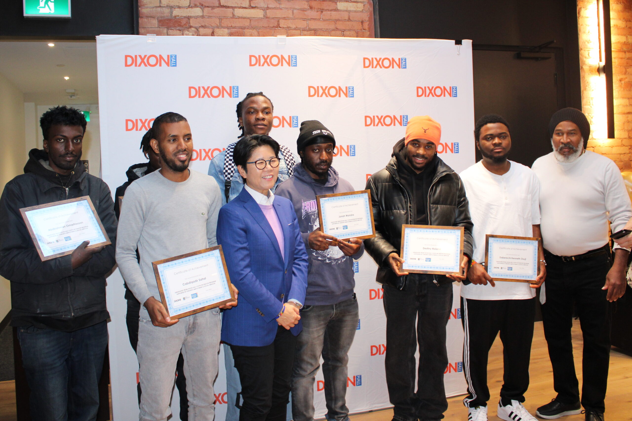 a-group-of-people-stands-in-front-of-a-dixon-hall-backdrop-holding-framed-certificates-they-are-smiling-and-posing-for-a-photo-dressed-in-casual-and-business-attire-the-setting-features-exposed-brick-walls-and-warm-lighting-