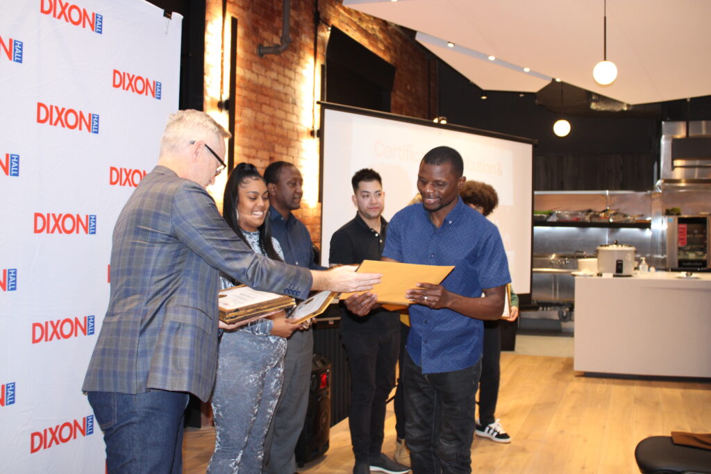 a-man-in-a-plaid-blazer-is-handing-a-certificate-and-envelope-to-a-smiling-recipient-in-a-blue-shirt-at-a-dixon-hall-event-other-attendees-and-organizers-stand-nearby-with-a-presentation-screen-and-kitchen-area-visible-in-the-background-