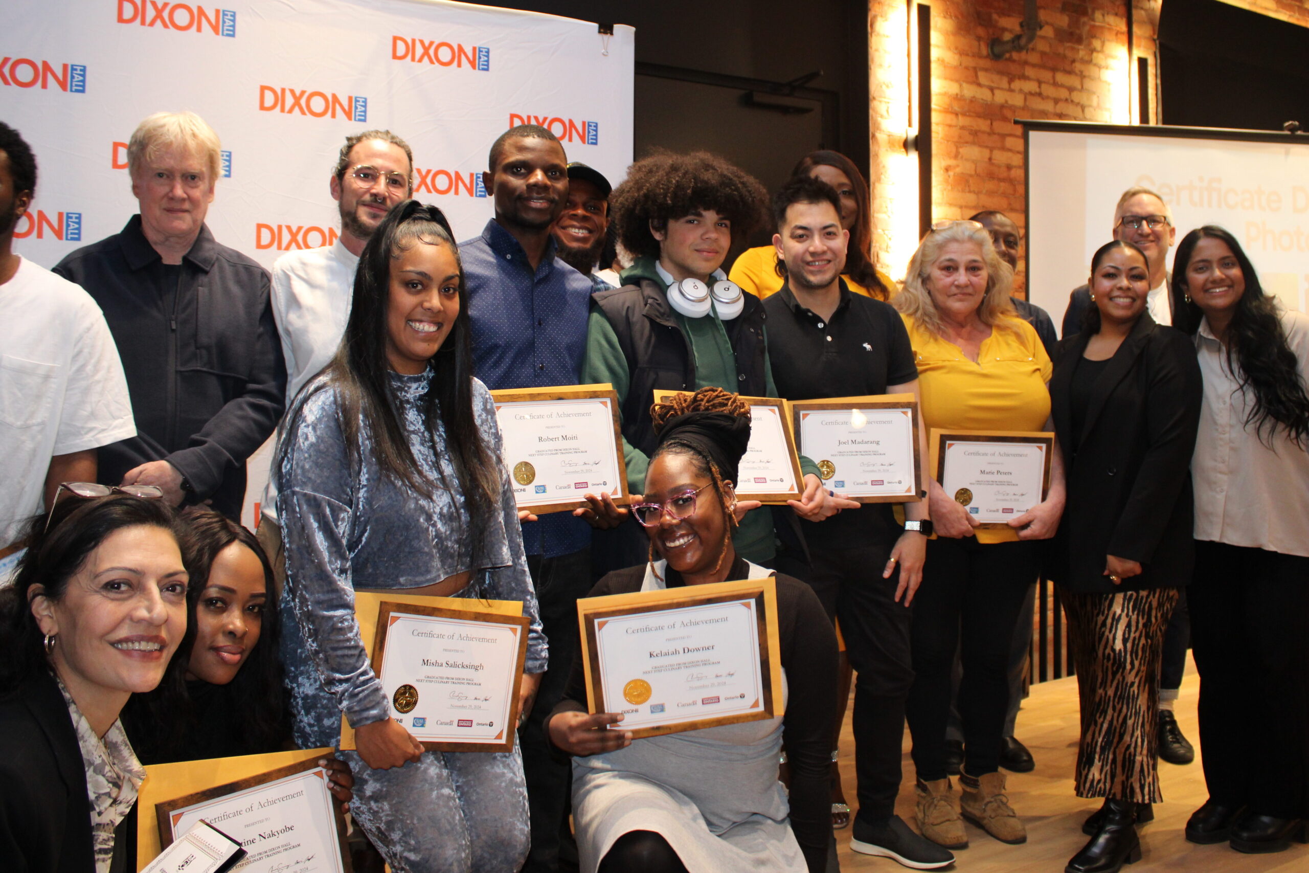 a-large-group-of-people-is-gathered-in-front-of-a-dixon-hall-backdrop-smiling-and-holding-framed-certificates-of-achievement-the-setting-has-exposed-brick-walls-and-warm-lighting-with-a-presentation-screen-visible-in-the-background-