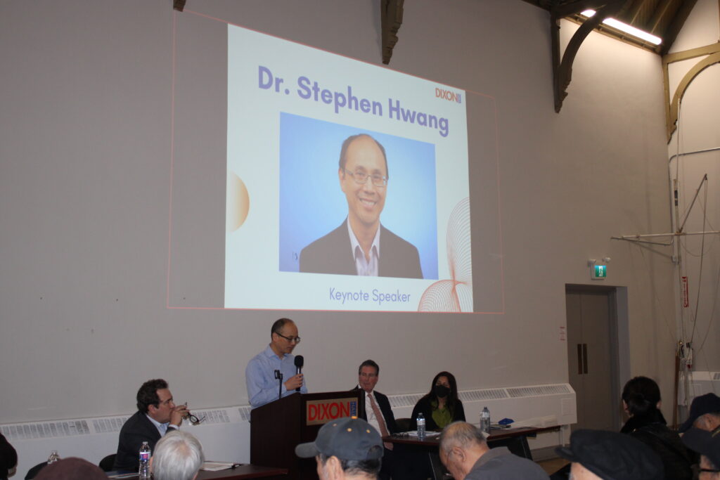 a-speaker-dr-stephen-hwang-is-addressing-an-audience-at-a-formal-event-a-large-screen-behind-him-displays-his-name-photo-and-title-as-the-keynote-speaker-panelists-are-seated-at-a-table-beside-him-and-attendees-are-listening-the-room-has-high-ceilings-and-a-professional-setting