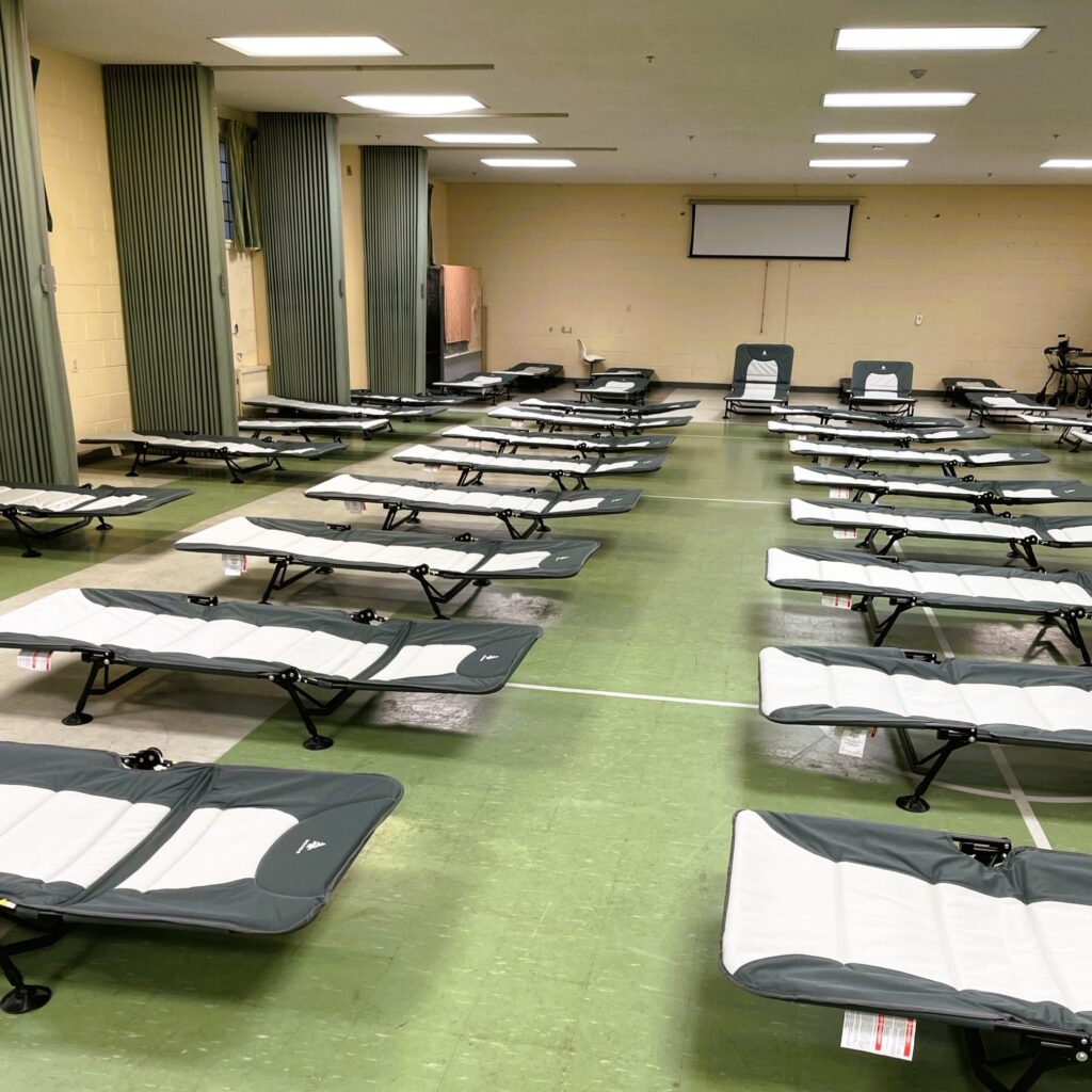 a-large-indoor-space-is-set-up-with-multiple-foldable-cots-arranged-in-rows-ready-for-use-the-room-has-green-flooring-beige-walls-and-a-projector-screen-at-the-front-curtains-cover-the-windows-along-one-side-